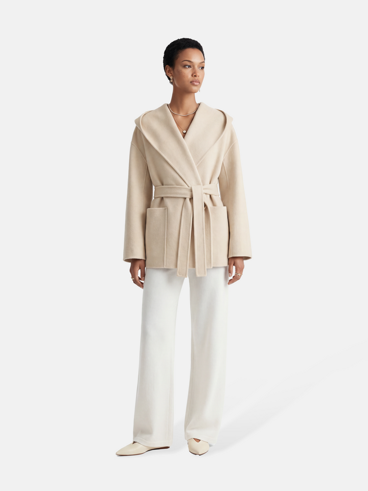 Full-length image of a cream wool belted overcoat with sculpted collar, self-tie belt, and oversized patch pockets, draped elegantly.