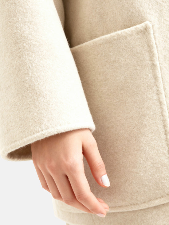 Full-length image of a cream wool belted overcoat with sculpted collar, self-tie belt, and oversized patch pockets, draped elegantly.